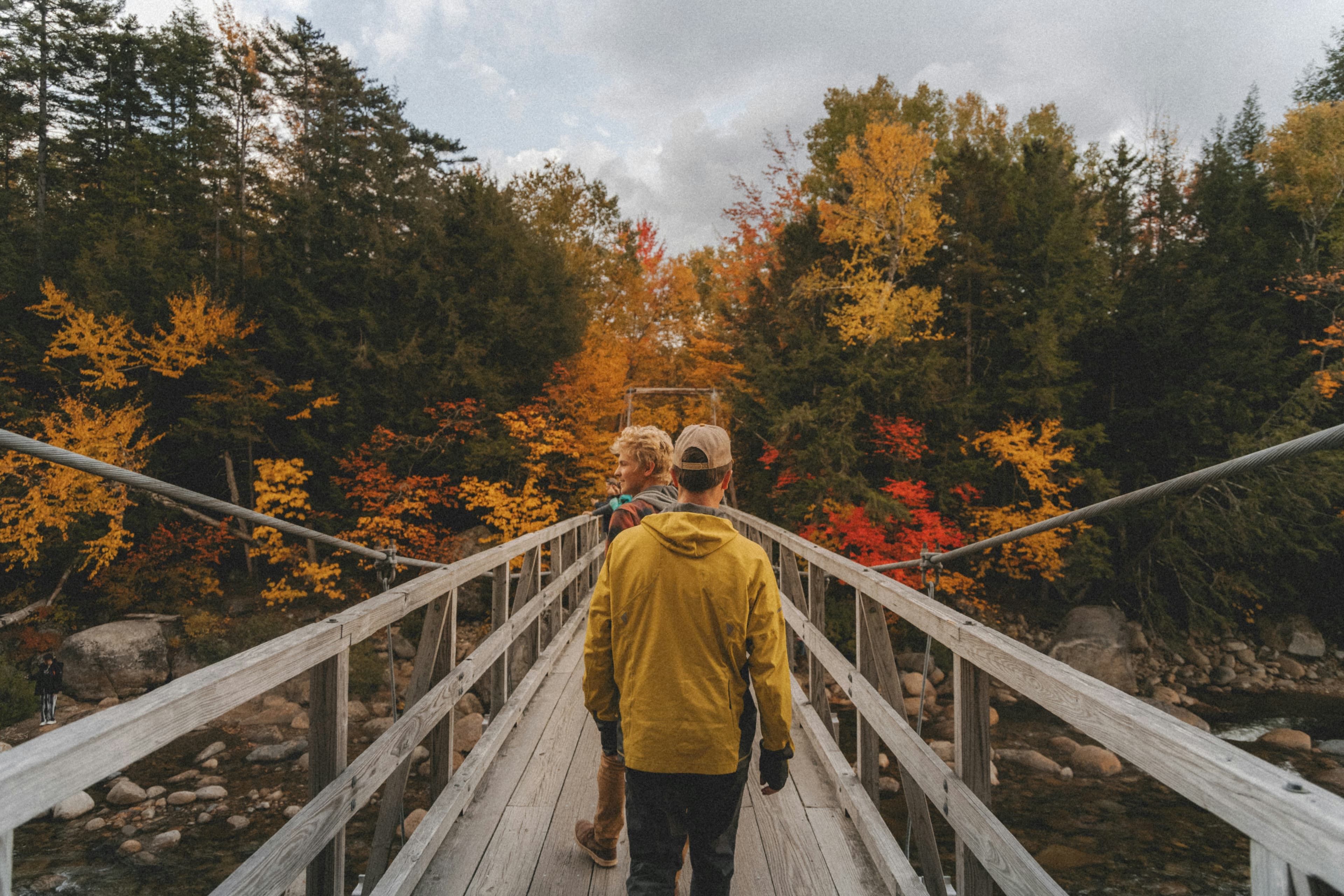 Two men walk across abridge towards a forest with fall colors